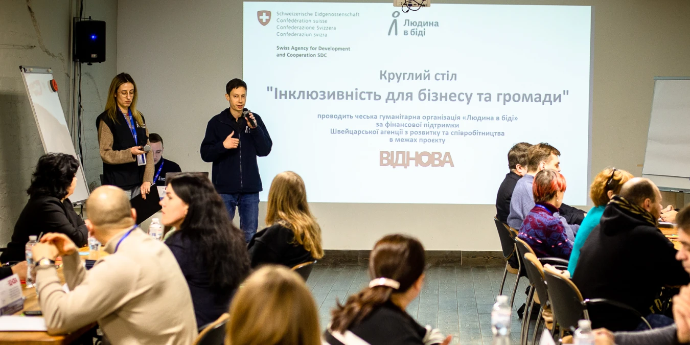 На зображенні показано круглий стіл на тему “Інклюзивність для бізнесу та громади”, який проводить гуманітарна організація “Людина в біді” за підтримки Швейцарської агенції з розвитку та співробітництва в межах проєкту “Віднова”. У центрі — спікер із мікрофоном, поруч стоїть представниця організації. У залі за столами сидять учасники заходу та слухають виступ. На екрані позаду демонструється офіційний слайд із логотипами та назвою події.