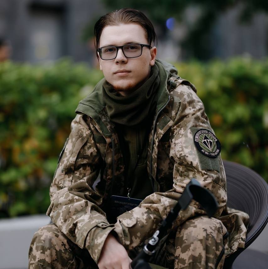 На рисунку зображено ветерана війни Андрія Голопапа. Він сидить на чорному стільці, одягнений у військову форму, у правій руці він тримає милицю з опорою під лікоть.
