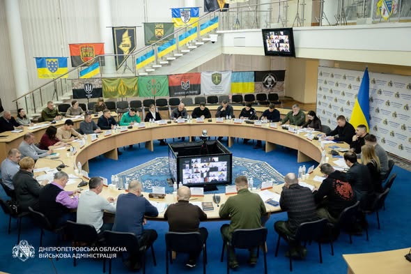 На рисунку зображено членів робочої групи “Прозорість та підзвітність” Дніпропетровської обласної військової адміністрації, які сидять за круглим столом. У середині круглого столу знаходиться чотирьох сторонній монітор, на якому видно інших учасників засідання робочої групи. Вони підключені віддалено за допомогою програмного забезпечення Zoom.
