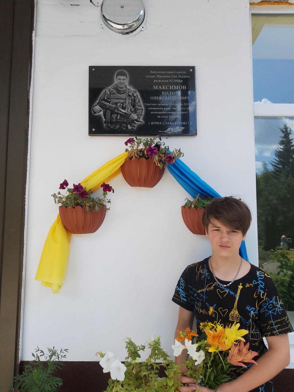 На рисунку зображено молодого хлопця, який стоїть на фоні пам'ятної таблички загиблого героя України Максимова Віктора Олександровича