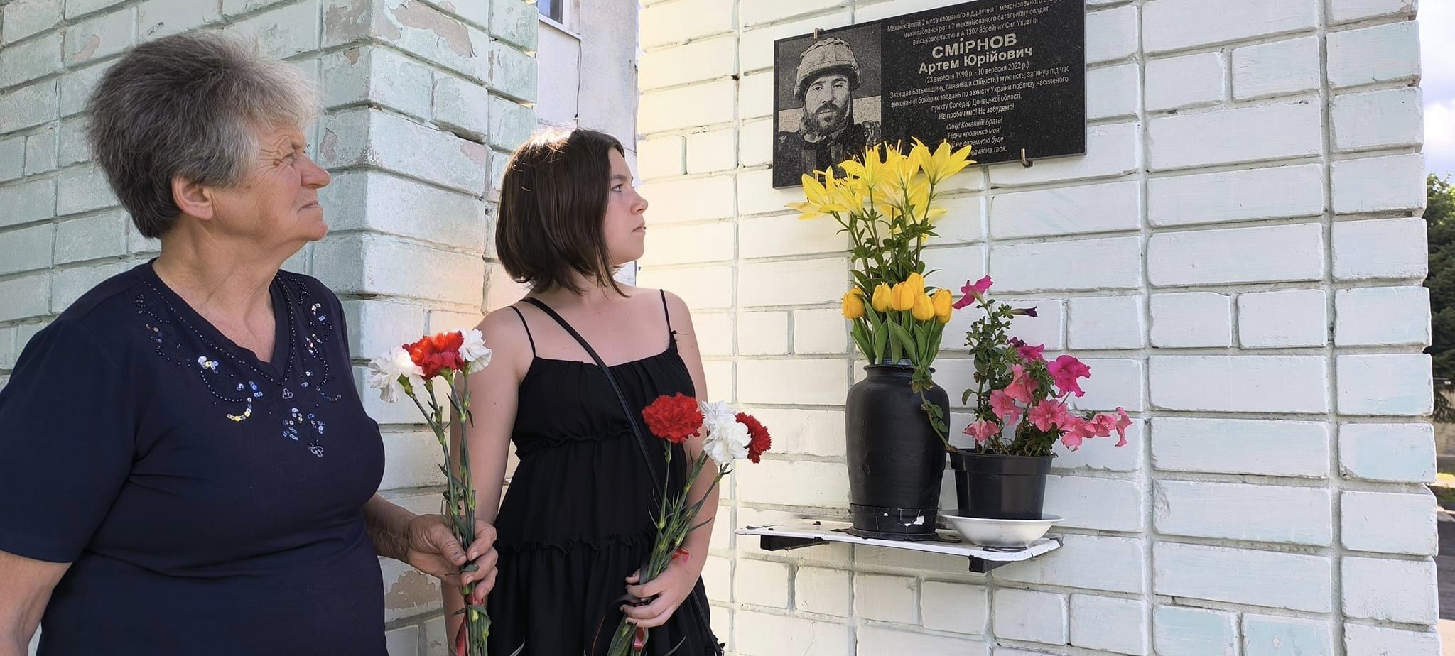 На рисунку зображено дві жінки з квітами (червоні рози), перша жінка похилого віку, друга молода. Вони стоять навпроти стіни пошани та пам'яті героям України, на якій весить пам'ятна табличка Смірнову Артему Юрійовичу