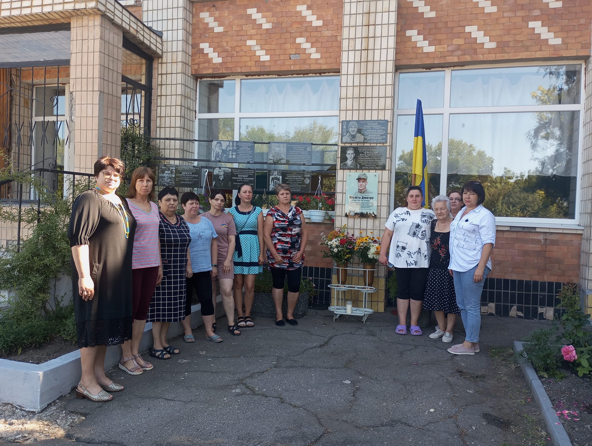 На рисунку зображено групу людей, які стоять на фоні школи. На стіні цієї школи зображено пам'ятні таблички загиблих героїв України, які навчалися у цій школі.