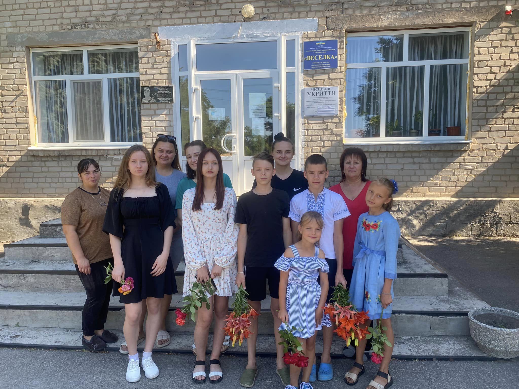 На рисунку зображено гурт дітей дитячого садка "Веселка" , тримаючих в руках квіти на фоні вище вказаного дитячого садка