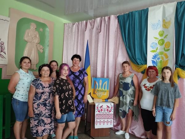 На рисунку зображено групу людей, які стоять на фоні прапора України, герба та рушника з традиційною червоною вишиванкою на честь святкування 28-ї річниці Конституції України.