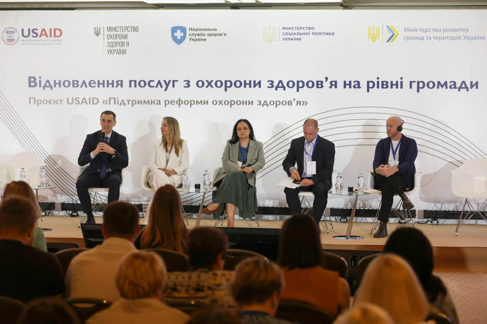 На рисунку зображено групу людей уважно слухаючих семінар на тему: "Відновлення послуг з охорони здоров'я на рівні громади". Проєкт USAID "Підтримка реформи охорони здоров'я". Також на рисунку зображено 5 менторів, які проводять дану конференцію.